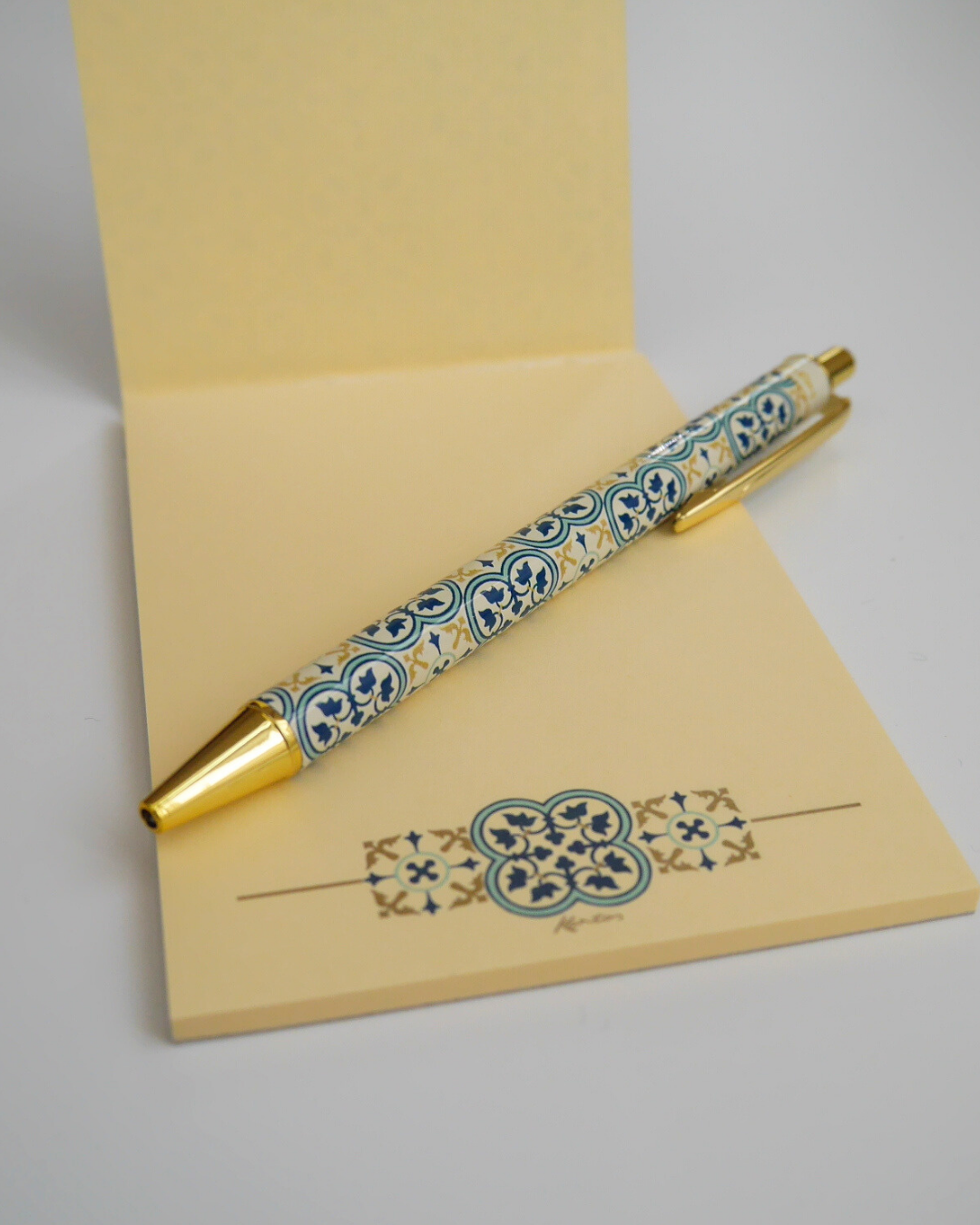 Quadrilobo柄のボールペンとメモ帳を合わせて置いたイメージ。金のパーツとクラシカルな模様が上品に映える。