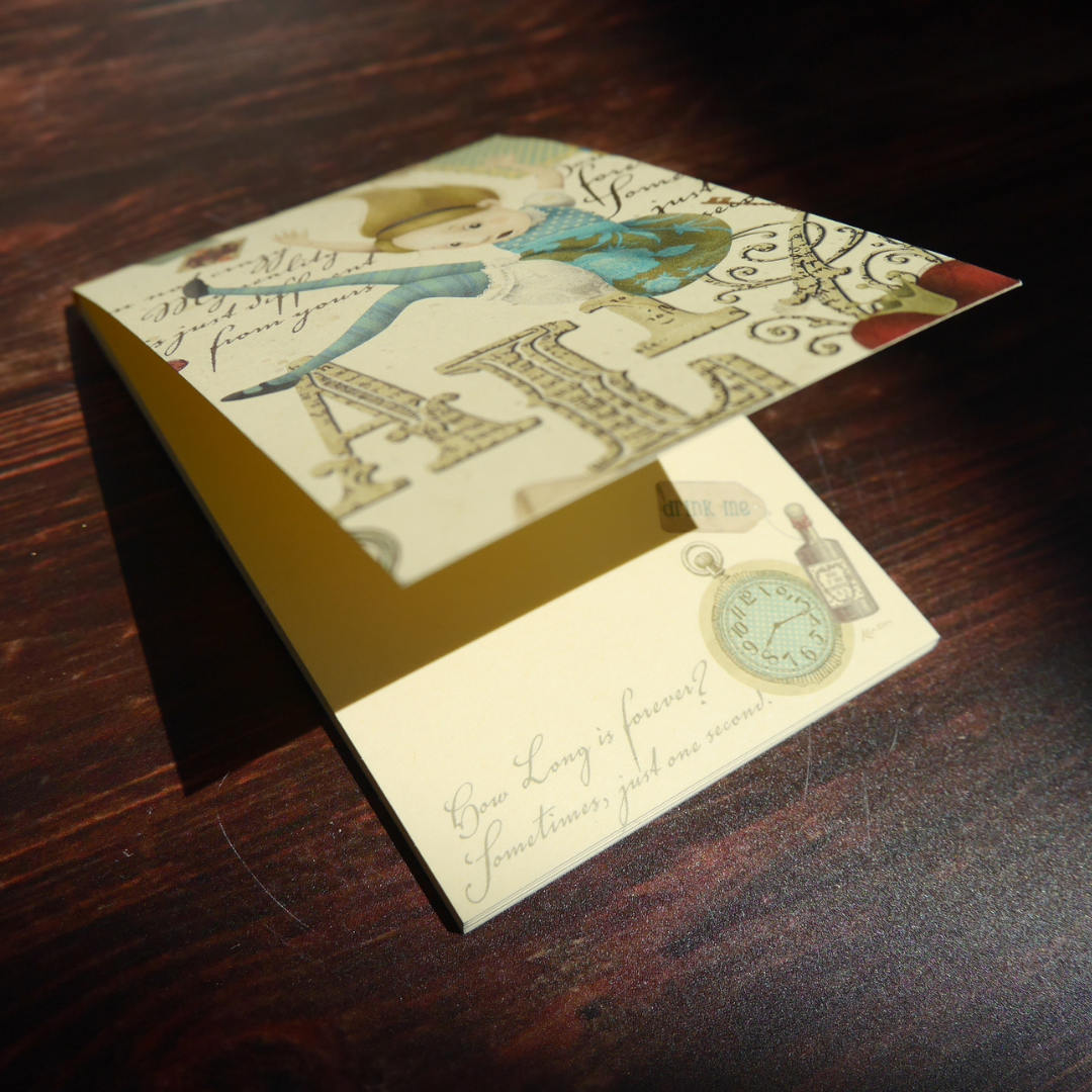 木の上に置かれたKartosのアリス柄メモ。光を受けて金粉が柔らかくきらめく、美しい紙面。